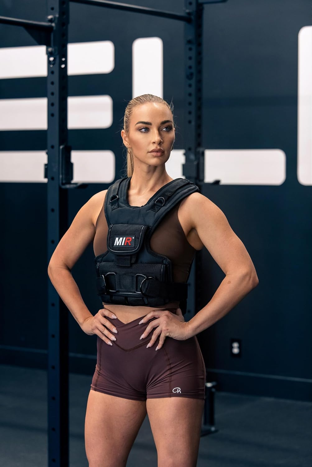 Woman standing with hands on hips wearing a black miR weighted vest in a gym