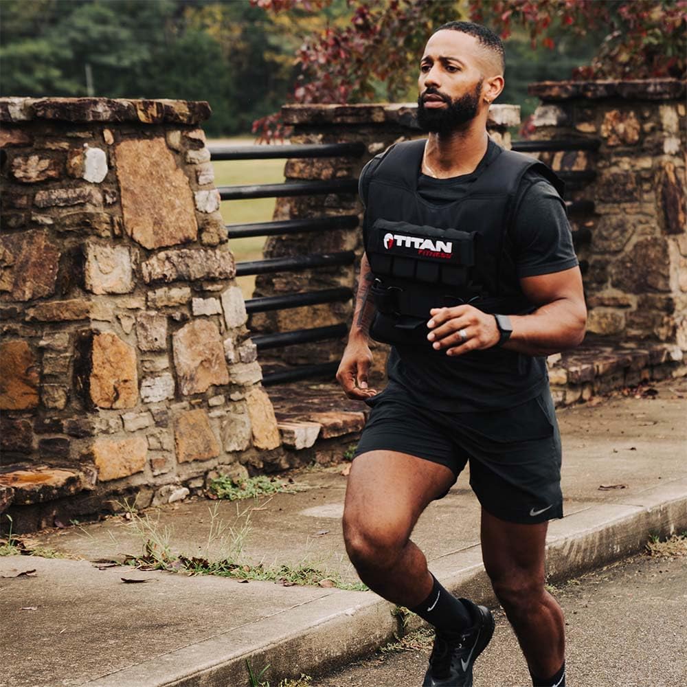 Person jogging outdoors beside a stone wall while wearing a black Titan Fitness weighted vest