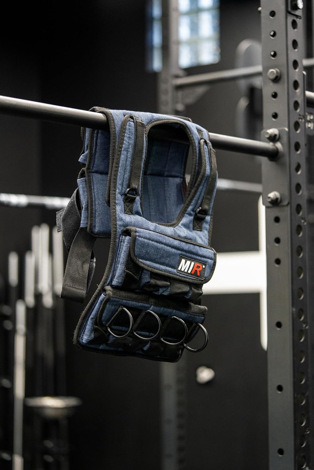Blue-gray miR weighted vest hanging from a gym rack bar with adjustable straps and front pockets visible